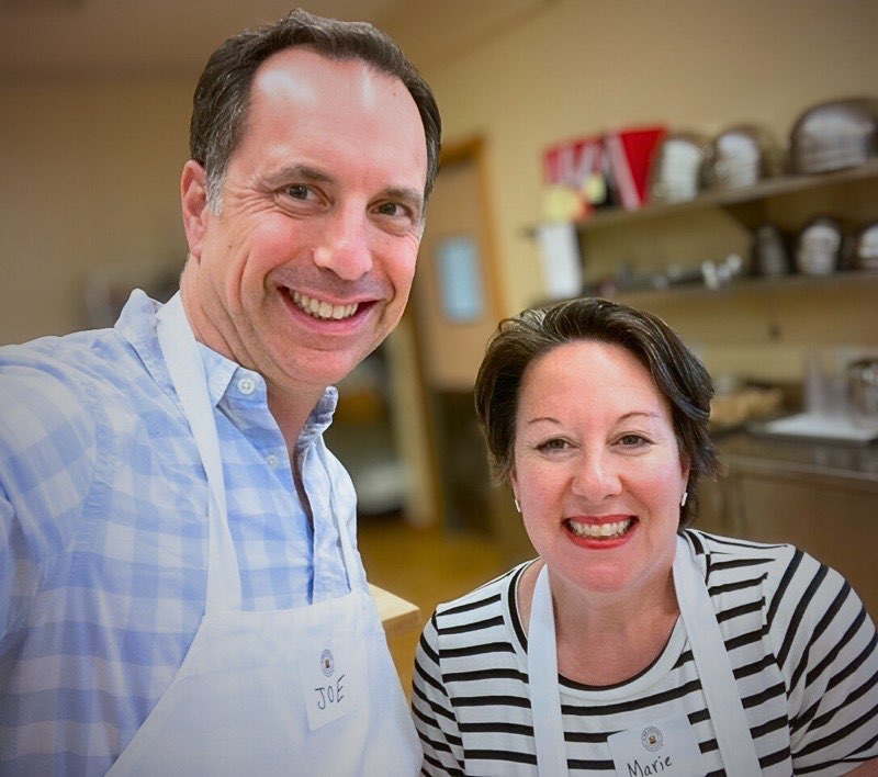 Joe and Marie Schick smiling in a teaching kitchen at a King Arthur baking class, both wearing white aprons.